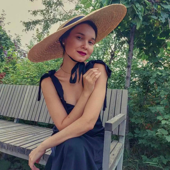 Woman sitting on a bench wearing a large wheat straw hat with a black ribbon under the chin