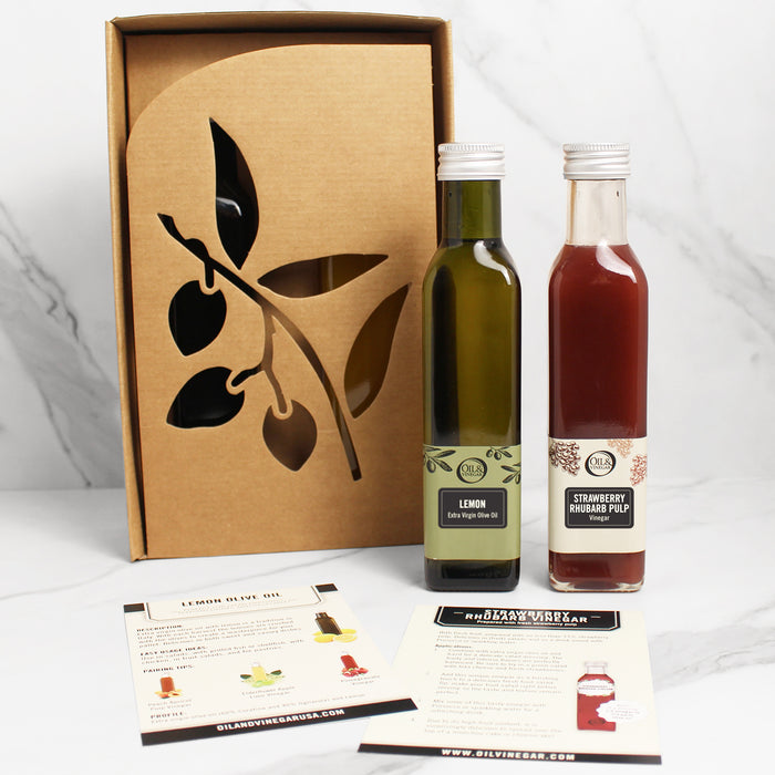 Two bottles of gourmet cooking oils and vinegars, one labeled Lemon Olive Oil and the other Strawberry Rhubarb Pulp Vinegar, presented next to a cardboard box with a printed leaf design.