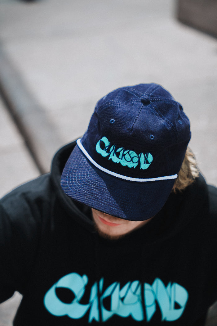 A person wearing a Quicksand Ink Blue Corduroy Hat and a black hoodie, both featuring matching turquoise abstract Quicksand logos, is seated outdoors on gray pavement.