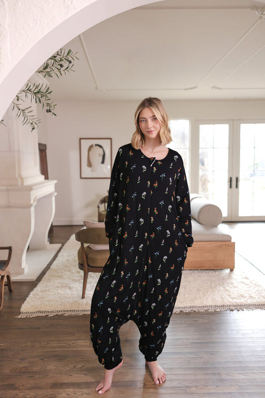 A woman stands barefoot in a bright, modern room wearing a black harem-style onesie with a small all-over floral print.