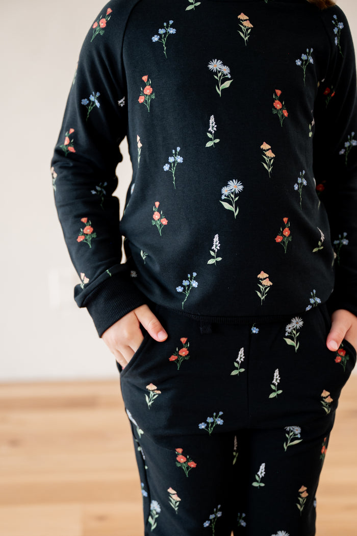 A person wearing a matching black long-sleeve top and pants set with a small multicolor floral print, standing indoors with hands in pockets.