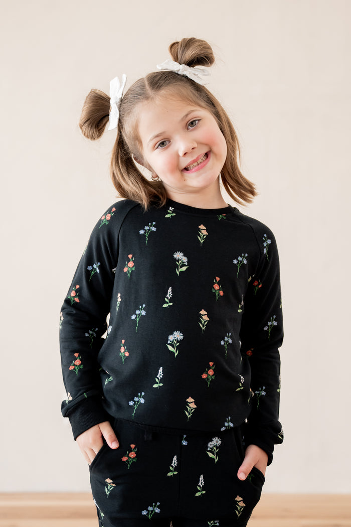 A young girl with brown hair in two buns wearing a black crewneck sweatshirt and matching sweatpants set, both decorated with a small multicolor floral pattern. She is smiling and posing with her hands in her pockets against a plain beige background.