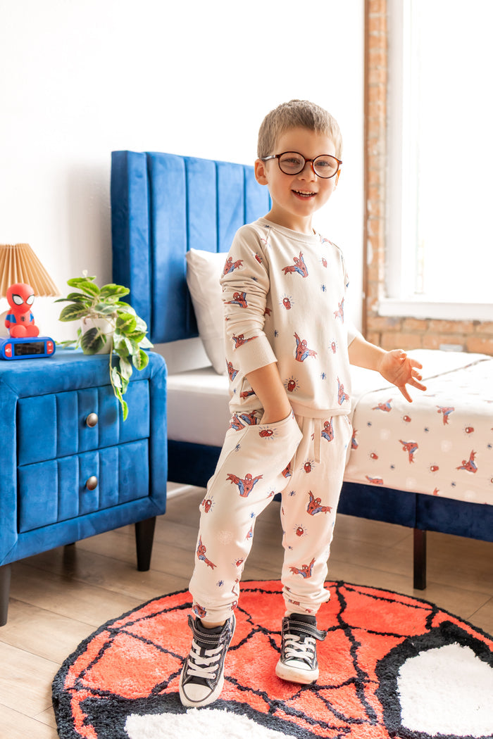Boy with specs standing on a spider man rug in a room with blue bed and side table wearing an oat color Long Sleeve Jogger Set with Spider-Man print