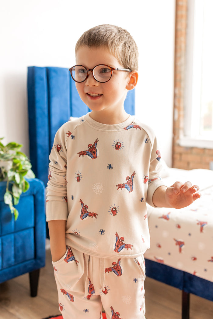 Boy with specs standing in a room with blue bed and side table wearing an oat color Long Sleeve Jogger Set with Spider-Man print