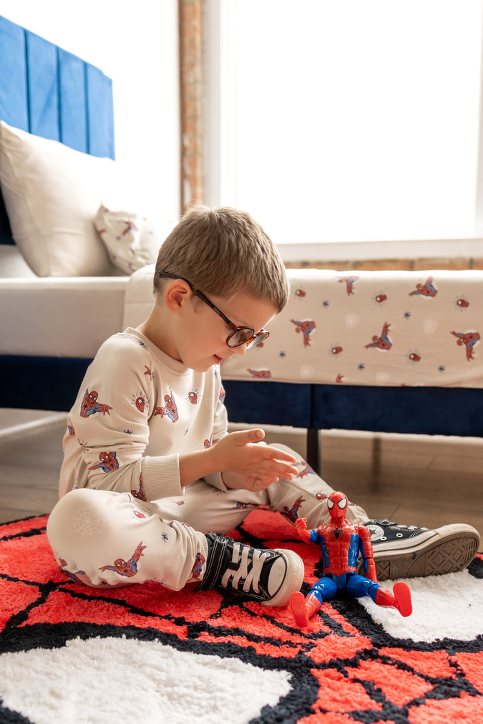 Boy with specs sitting on a spider man rug and spider man toy in a room with bed wearing an oat color Long Sleeve Jogger Set with Spider-Man print