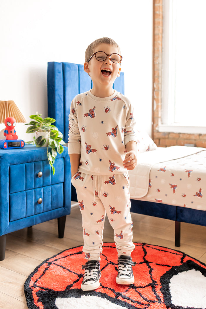 Boy with specs smiling on a spider man rug in a room with blue bed and side table wearing an oat color Long Sleeve Jogger Set with Spider-Man print