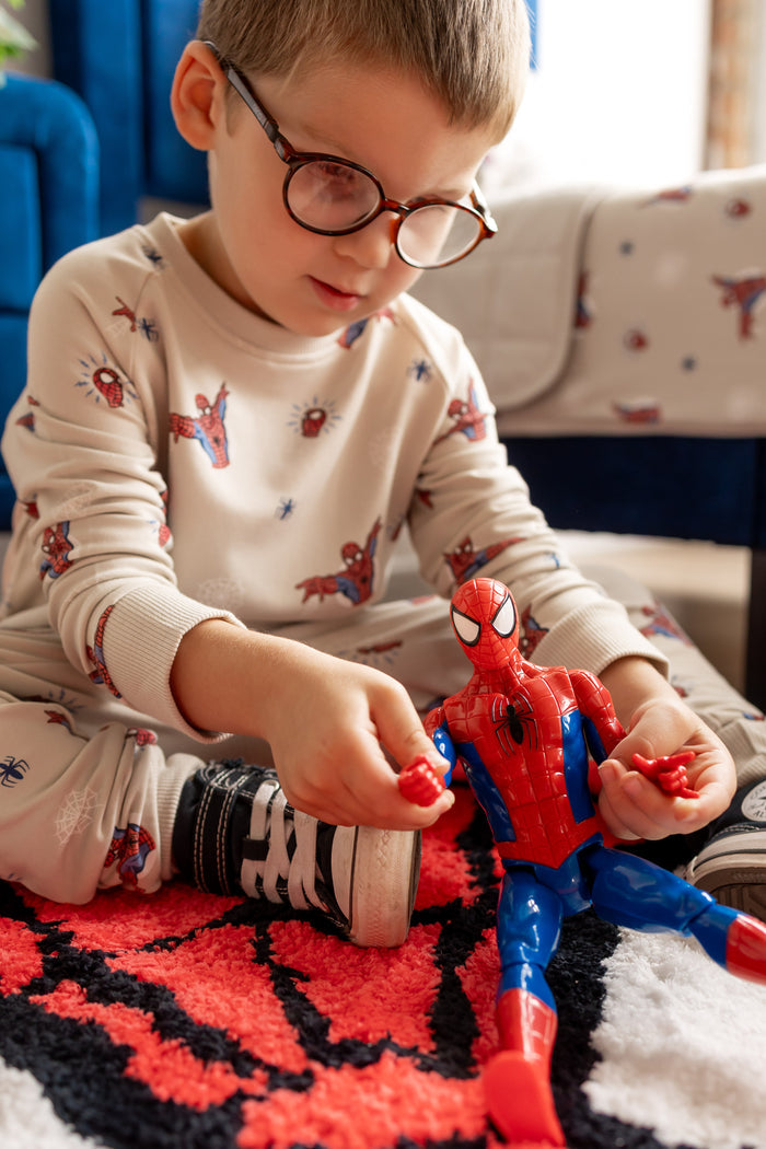 Boy with specs playing with spider man toy sitting on a spider man rug in a room wearing an oat color Long Sleeve Jogger Set with Spider-Man print
