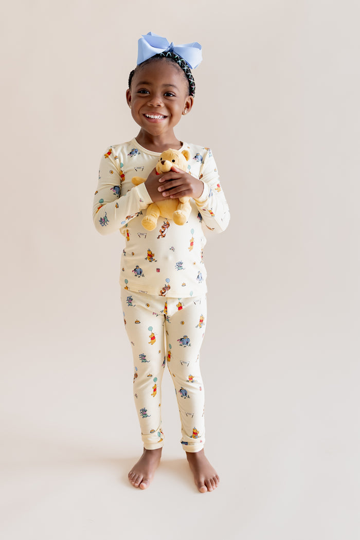 Girl smiling in Long Sleeve Pajamas in Winnie the Pooh Celebration print holding stuffed toy on a beige background