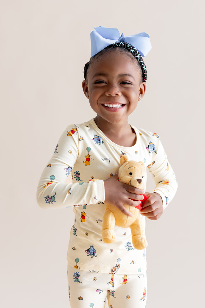 Girl wearing Long Sleeve Pajamas in Winnie the Pooh Celebration print holding stuffed toy on a beige background