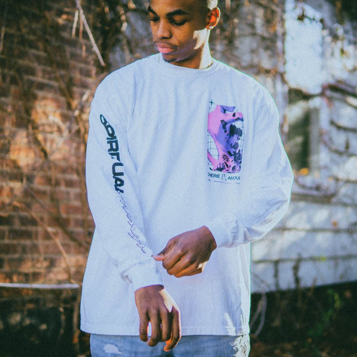 Someone wearing White longsleeve shirt with SPIRITUAL written in black letters down the right sleeve. In the top left corner of the shirt, there is a design of a woman with tattoos on her back. The woman is colored pink and purple with black tattoos, and below reads CHERIE AMOUR.