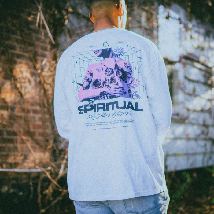 Someone wearing White longsleeve shirt with SPIRITUAL written in black letters down the right sleeve. In the top left corner of the shirt, there is a design of a woman with tattoos on her back. The woman is colored pink and purple with black tattoos, and below reads CHERIE AMOUR.