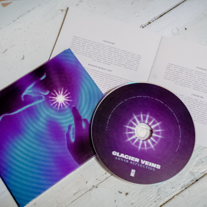 Square CD with a drawing of two hands reaching for a star. Around the star and covering the whole album cover is a ripple effect drawn in purple and green. To the right of the cover is a disc that is purple and says GLACIER VEINS on the bottom of the disc. There is a star bursting out of the center of the disc. Both are laying on top of a lightly colored wooden surface.