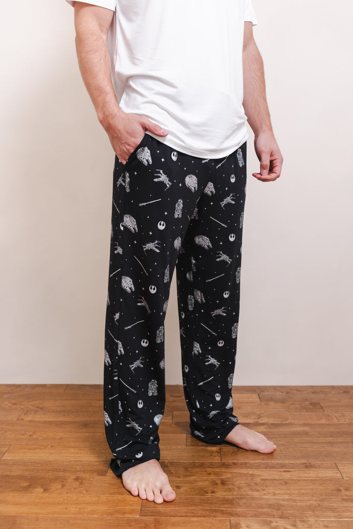 A person wearing black pajama pants with a white space-themed print of planets, stars, and Rebel Alliance symbols, paired with a white t-shirt, standing on a wooden floor against a plain beige wall.