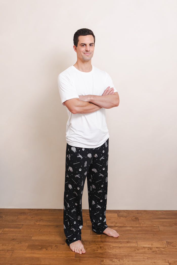 A man standing barefoot on a wooden floor against a plain beige wall, wearing a white short-sleeve t-shirt and black pajama pants with a white Star Wars pattern.