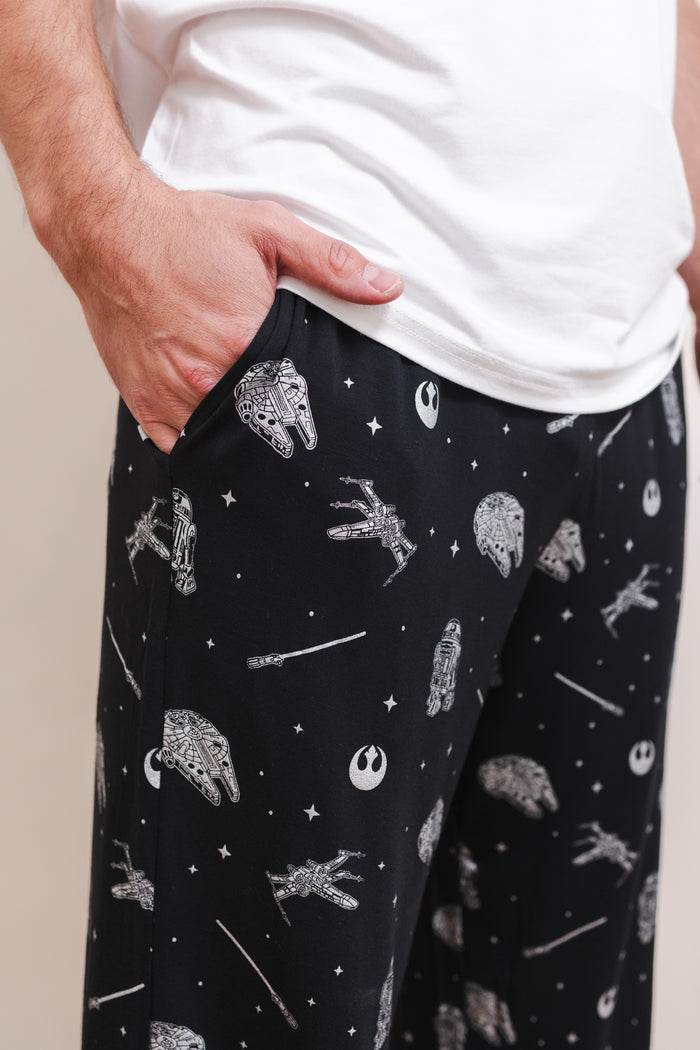 A close-up of a person wearing black pajama pants with a white space-themed print featuring Rebel Alliance symbols, stars, and spacecraft, paired with a white t-shirt.