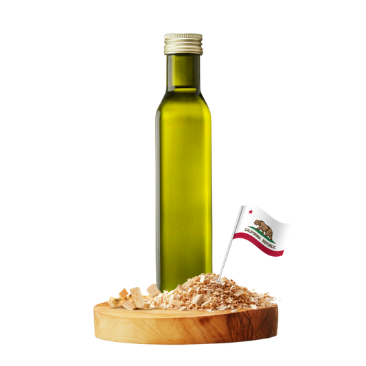 A tall olive-green glass bottle with a silver cap stands on a round wooden board with several wood chips and a small California flag.