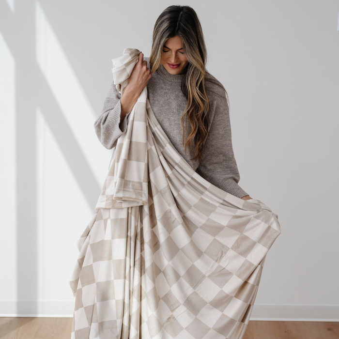 A woman is holding a tan and cream checkered minky stretch blanket. This soft blanket is Saranoni blanket that is very stretchy and durable. Use this throw blanket for lounging, gifting, or on the go.