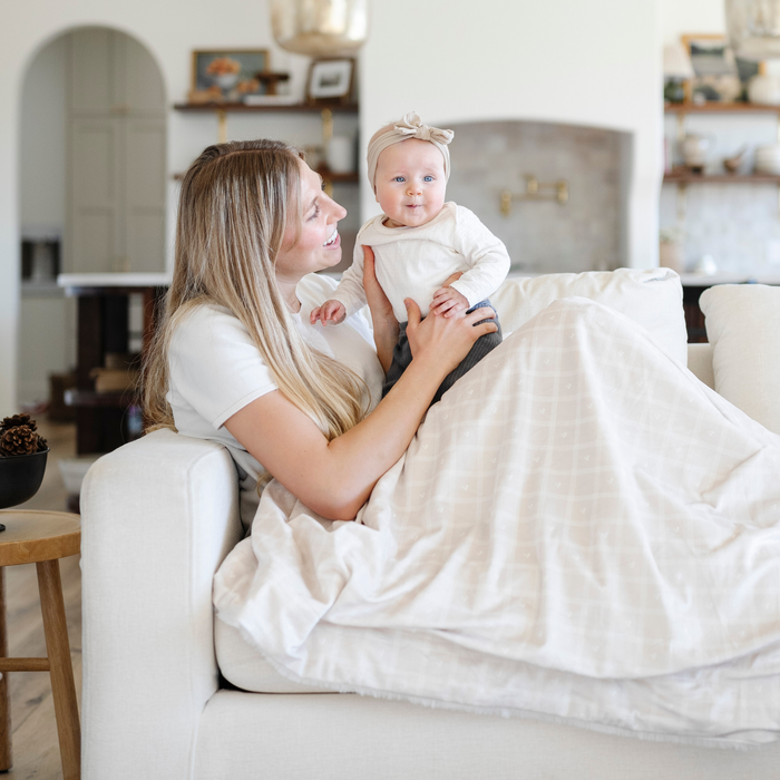 A mother holds her baby girl and is sitting with a luxuriously soft Saranoni minky/lush blanket. This premium dual-textured extra-large throw features Saranoni’s signature cream lush fabric on one side and ultra-soft square printed minky on the other. Designed for ultimate relaxation, this plush blanket is perfect for snuggling, movie nights, and adding a touch of luxury to any home. This soft blanket is a luxury blanket and a Saranoni blanket. The cozy blanket is a comfortable blanket.