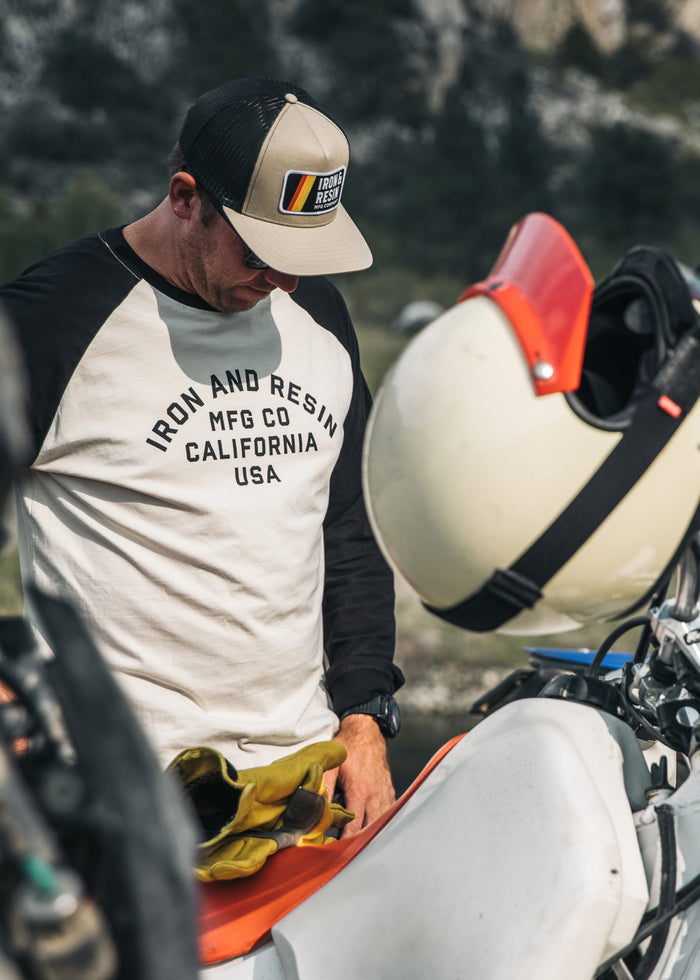 Man in Iron and Resin shirt and cap stands by a motorcycle, holding gloves. Helmet rests on the bike, outdoors.