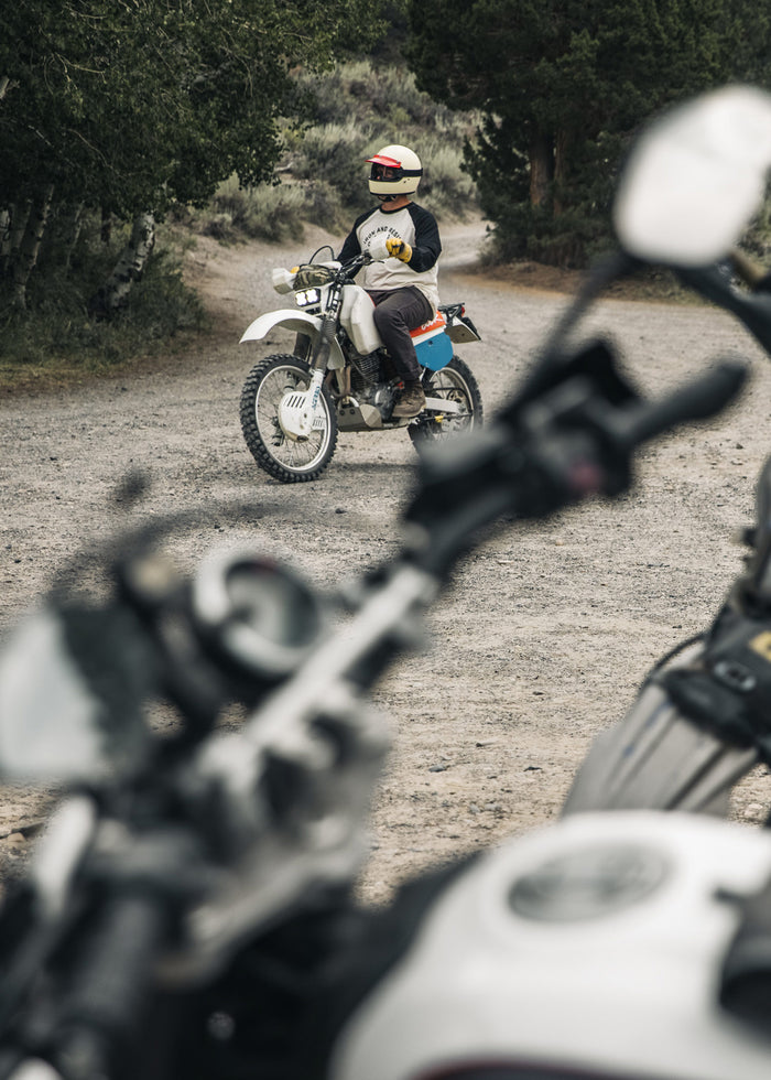 A motorcyclist in a helmet pauses on a dirt path, surrounded by trees, with another bike blurred in the foreground.