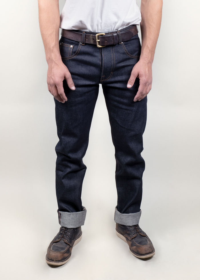 Man in dark jeans and brown leather shoes stands against a plain background, showcasing casual style with a white shirt.