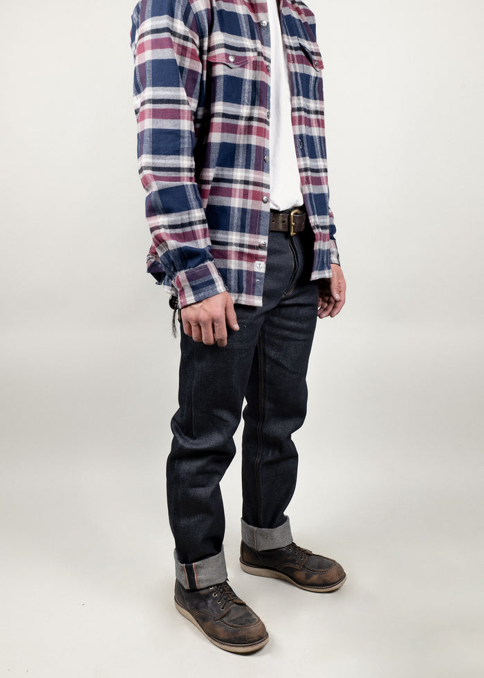 Man in plaid shirt, white tee, dark jeans, and brown boots stands against a plain background, showcasing casual fashion style.