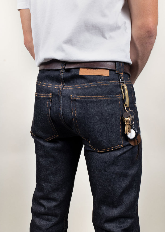 Man in dark jeans with a leather belt and keychain, wearing a white shirt, stands with hands behind back against a light background.