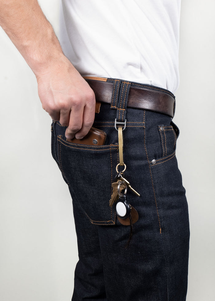Man in jeans and white shirt holds phone in pocket, keys with leather fob hang from belt loop.