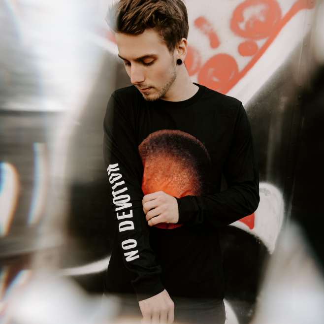 A young man with gauged ears adjusts his black No Devotion long-sleeve tee, which features NO DEVOTION on the sleeve and a bold red graphic on the front, standing before a graffiti-covered wall.