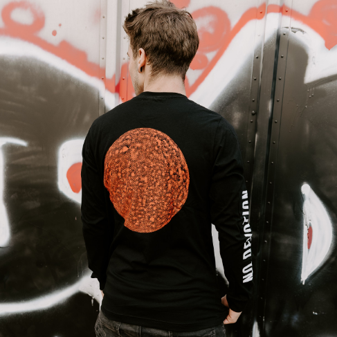 A person faces away from the camera, wearing a black No Devotion long-sleeve T-shirt with a large orange textured circle and "No Devotion" text on the right sleeve, standing in front of graffiti-covered metal.