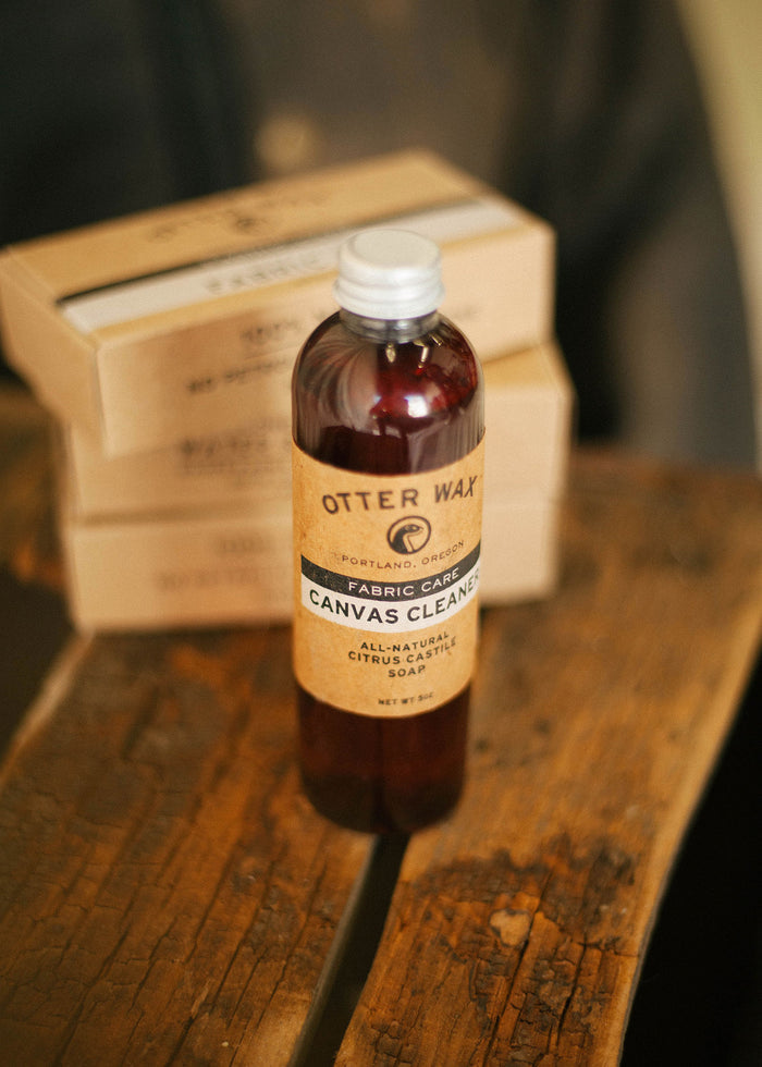 Otter Wax Canvas Cleaner bottle on rustic wood table, with boxes in the background, emphasizing fabric care and natural ingredients.