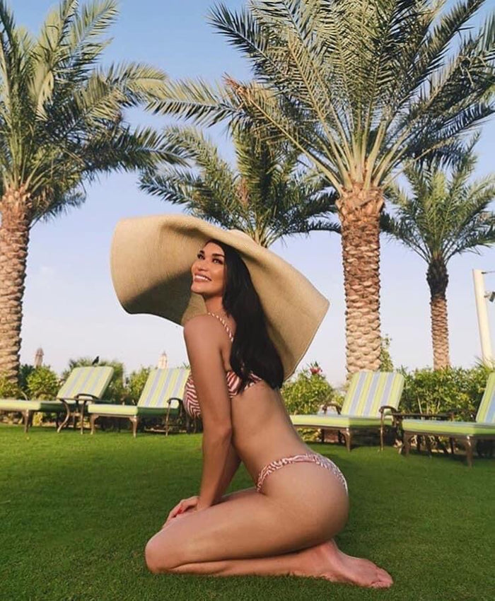 Woman wearing oversized straw sun hat kneeling on grass with palm trees and lounge chairs in background