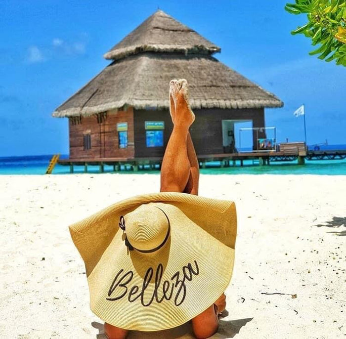 Person relaxing on beach wearing oversized straw sun hat with Belleza text near tropical water bungalow