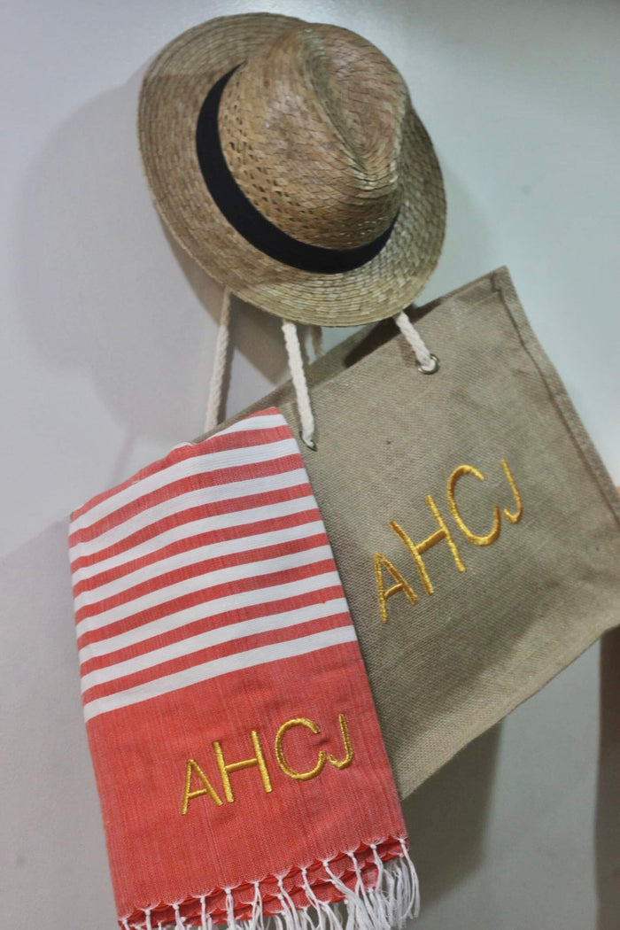 Personalized beach towel with red stripes and initials AHCJ hanging next to a straw hat and beige tote bag