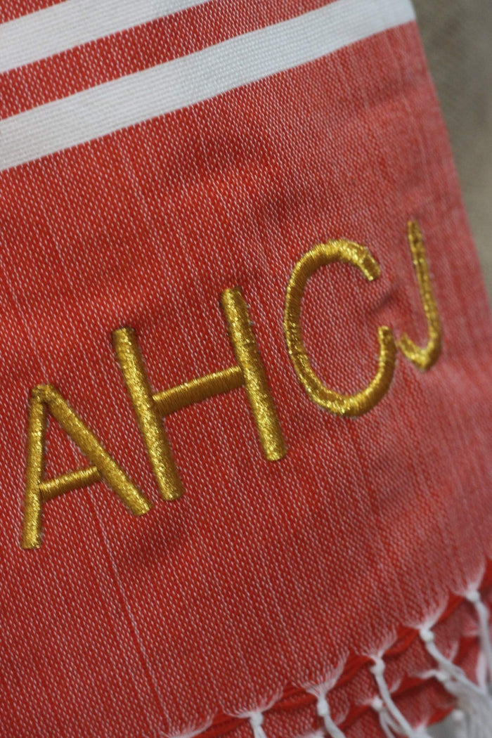 Personalized beach towel in red with gold embroidered initials and white striped edge from Travellers Hat Boutique