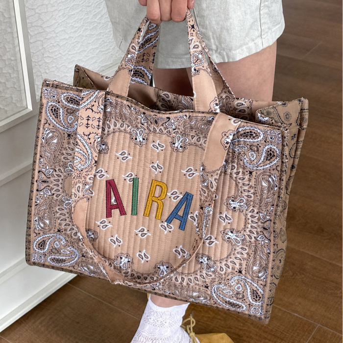 A person holds a large tan quilted tote bag with a paisley print and colorful embroidered letters spelling 'AIRA' on the front, shown in an indoor setting with wood flooring.