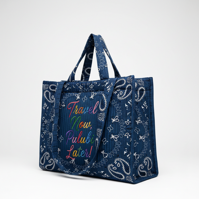 A navy blue tote bag with a white paisley bandana print and colorful embroidered text reading 'Travel Now, Pulubi Later!' on the front, shown against a plain white background.