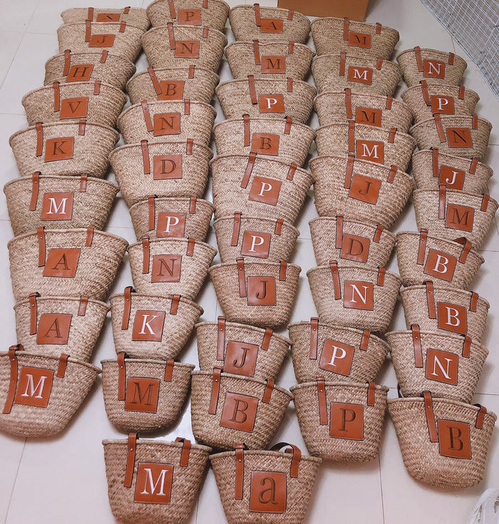 Multiple seagrass basket handbags with leather straps and initial patches arranged in rows from Travellers Hat Boutique