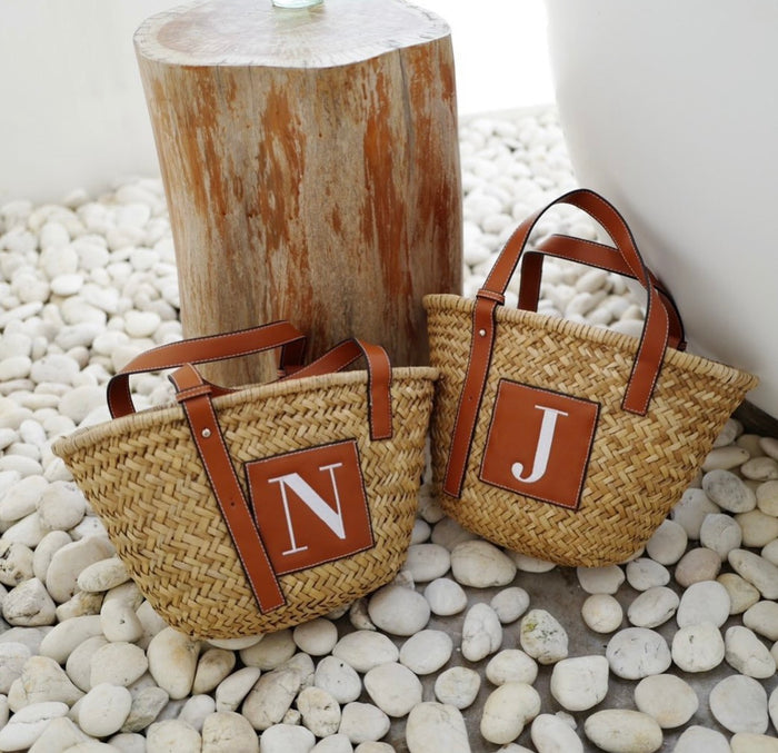 Seagrass basket handbag tote with brown leather handles and initial patches on white pebble ground