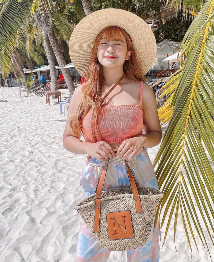 Woman on beach holding seagrass basket handbag with leather straps and monogram detail