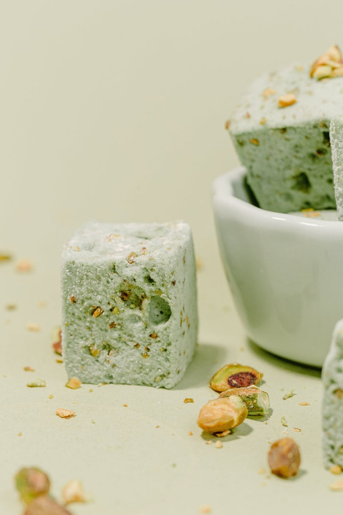 A close up shot of a XO Marshmallow gourmet pistachio marshmallow on its side. Next to it is half of a bowl with more marshmallows coming out of it. Pistachios are scattered across the photo all against a light mint green background.