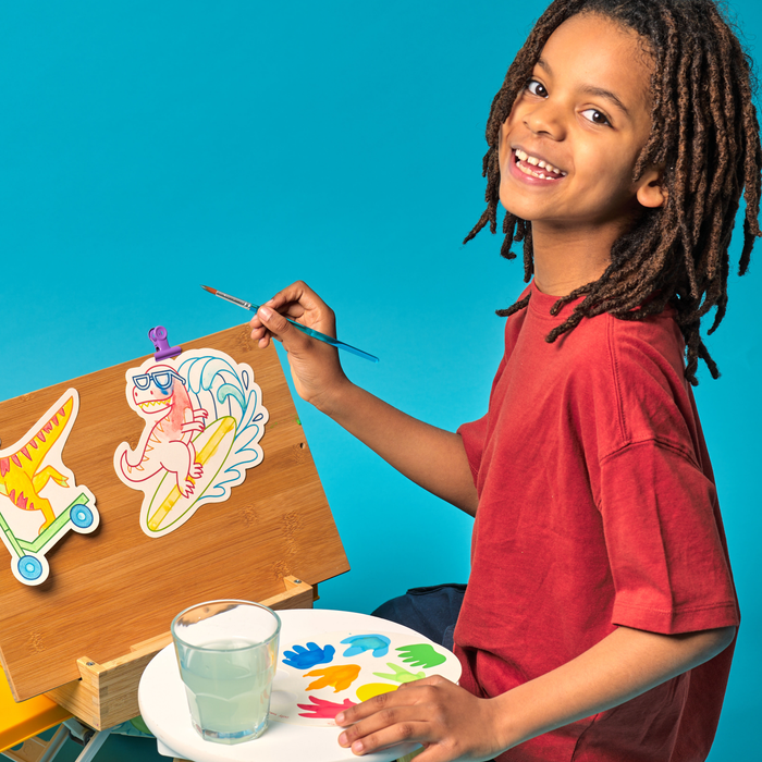 Kid smiling while painting dinosaur paint cards from the OOLY Chills and Thrills Dinos Playful Painters Easy Watercolor Painting Kit on a blue background