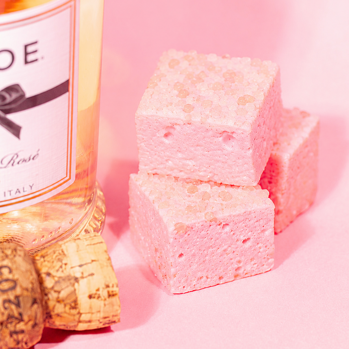 Pink square marshmallows pieces stacked next to a bottle of rose wine and corks on a pink background.