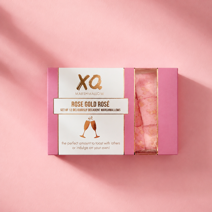 A pink rectangular box of XQ Marshmallow 'Rose Gold' decadent marshmallows, shown open to reveal pink marshmallows inside, set against a matching pink background with soft diagonal shadows.