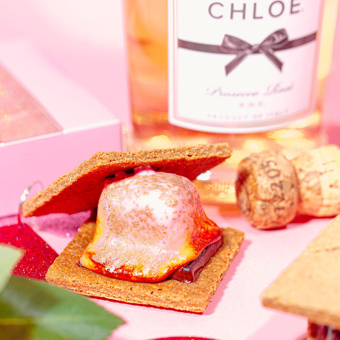 A styled close-up of a pink-themed scene featuring a bottle of Chloe Pinot Noir wine, corks, and a s'mores dessert with graham crackers and marshmallow, set against a pink background with decorative red glitter and flowers.