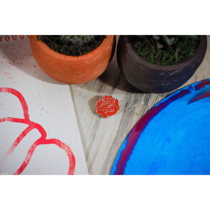 Pin of a small red rose. Pin is sitting on a wooden surface near plant holders.