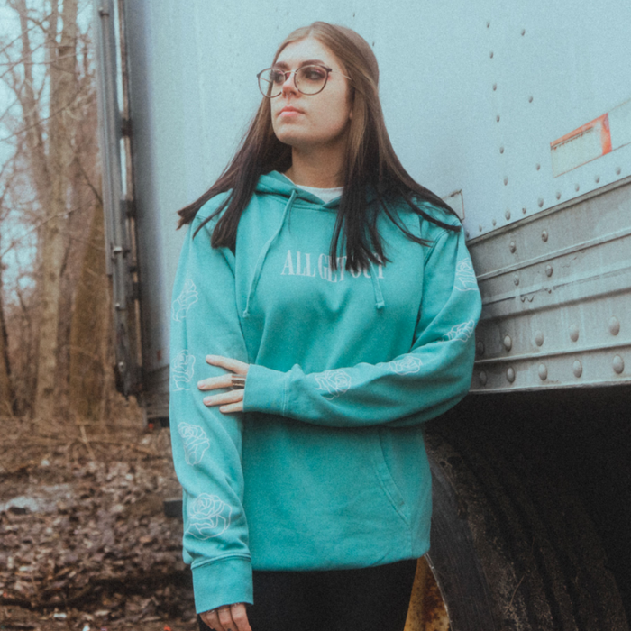 We see the front side of a soft green mint colored hoodie with text in the middle that reads, ALL GET OUT. On both sides of the sleeves we see roses depicted going all the way from the top of the outer arm to the bottom of the outer arm. The hoodie looks very comfortable and soft.