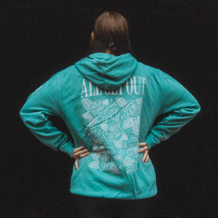We see the backside of a soft green mint colored hoodie with text on top that reads, ALL GET OUT. Below the text is an elegant display of roses that are shown as a white outline against the mint green hoodie color.