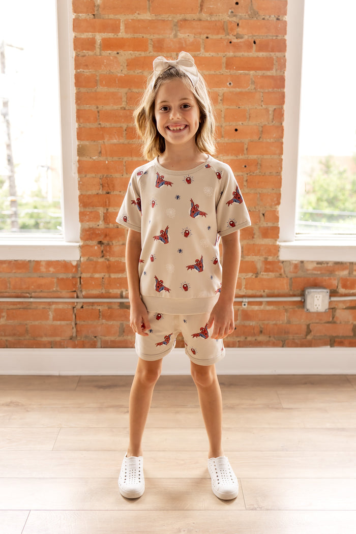Girl smiling in front of a brick wall wearing an oat color Short Sleeve Jogger Set with Spider-Man print with white shoes and oat color bow on head