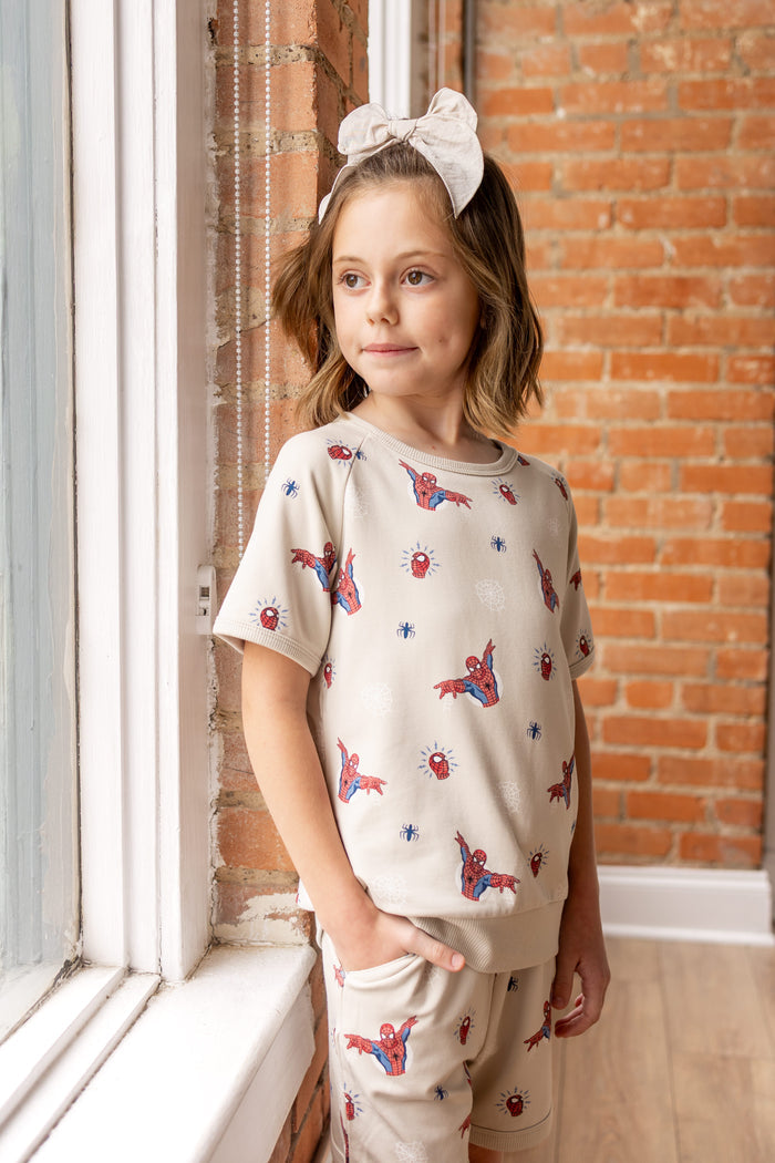 Girl standing in front of a brick wall against a window wearing an oat color Short Sleeve Jogger Set with Spider-Man print and oat color bow on head